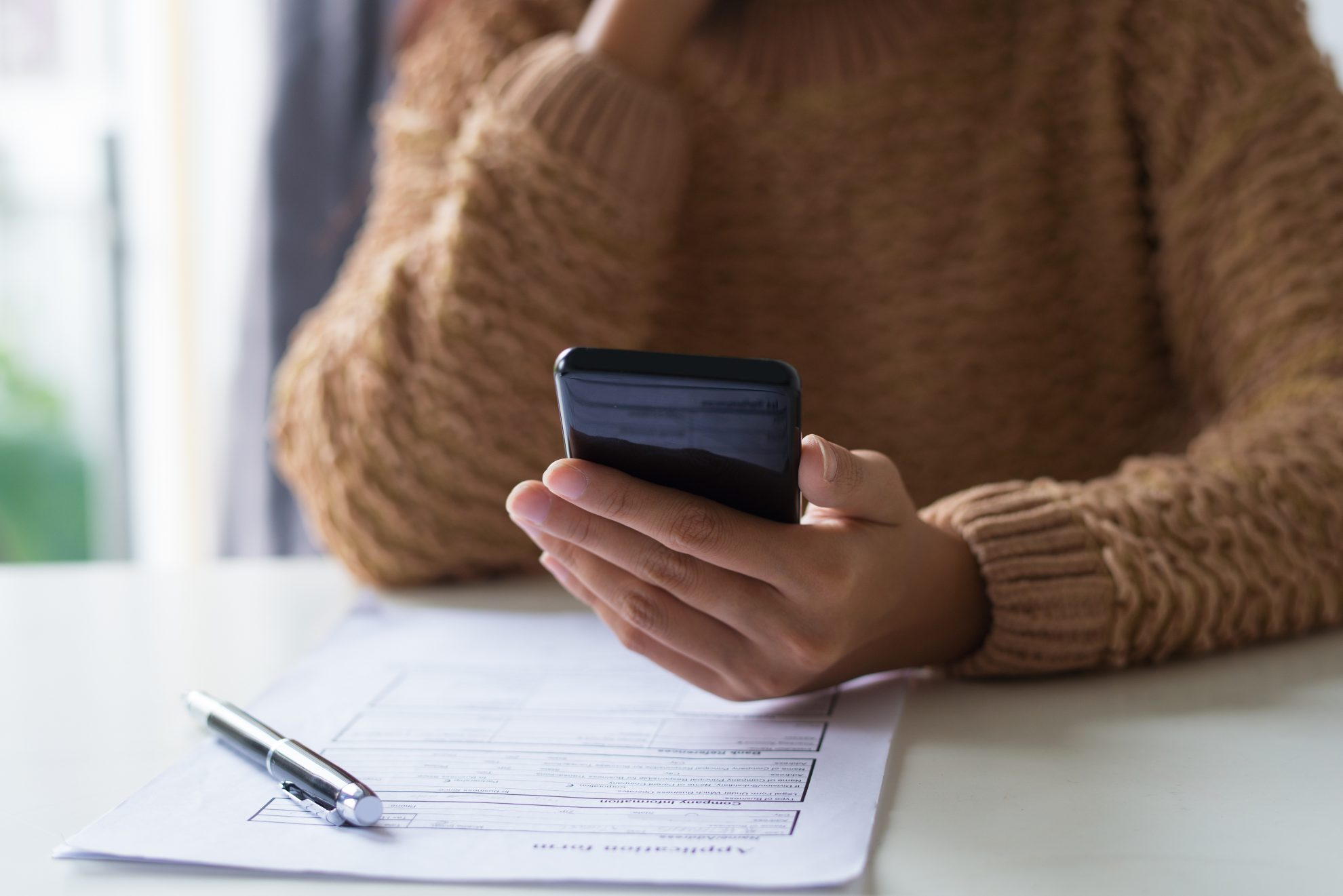 Close-up of busy lady filling document and checking sms on phone. Unrecognizable woman in warm sweater sitting at table and using smartphone. Work concept